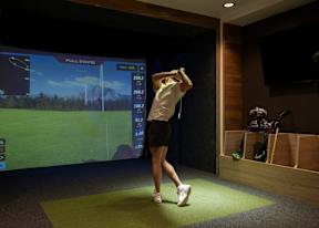A woman is playing golf in a room with a screen showing a golf course and her swing.