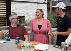 A woman in a pink dress is laughing while two men sit at a table with food and drinks.