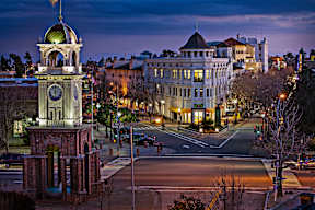 A clock tower stands in front of a building with a street in front of it.