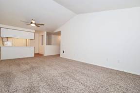 the spacious living room and kitchen of an apartment with carpeting and a ceiling fan