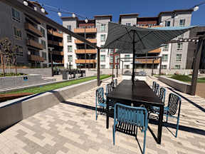 A black table with blue chairs and a black umbrella in front of apartment buildings.