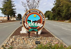 A welcome sign for the City of Scotts Valley is displayed in front of a road.