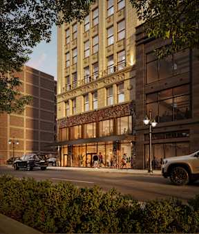 An exterior rendering of The Belle with a glass facade is illuminated from within, with cars parked in front and people walking on the sidewalk.