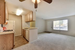 Vacant apatemtn home showing kitchen, living room and entry.  There is plush carpeting in living room and hardwood inspired flooring in the kitchen area. 