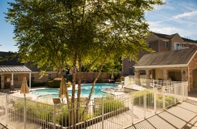 A gated sparkling swimming pool with a hot tub surrounded by plants, trees, tables with chairs and umbrellas near three-story apartment buildings.