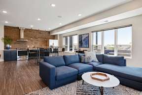 Fridley, MN Pet-Friendly Luxury Apartments - Axle Apartments - A Living Area Featuring A Navy Sectional Sofa, Large Windows, Round Coffee Table, Potted Plant, And Wood-Style Flooring.