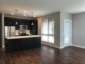 A kitchen with black cabinets, stainless steel appliances, pendant and recessed lighting, a window, and hardwood-style flooring with open access to the living room  at Keva Flats, Exton, 19341.