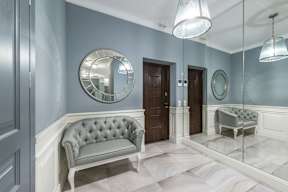A grey tufted sofa sits in a hallway with a mirrored wall and a round mirror above it.
