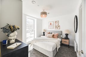 A bedroom with a large bed, a nightstand with a lamp, and a dresser with a vase of flowers on top.