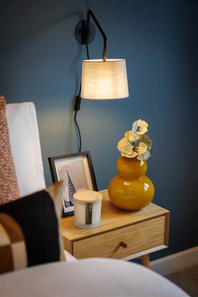 A yellow vase with flowers sits on a wooden nightstand next to a picture frame and a candle.