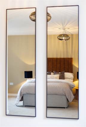 A bedroom with a bed, a lamp, and a chandelier is reflected in a large mirror.