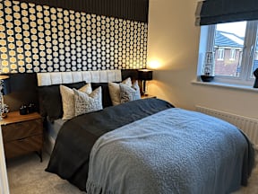 A bedroom with a black and white patterned wallpaper, a bed with grey bedding, and a window with a view of a red brick building outside.