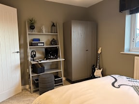 A room with a bed, a shelf with electronics, and a guitar.