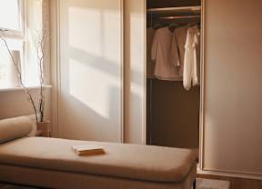 A bedroom with a bed, a window, a plant, a book, and a closet with clothes.