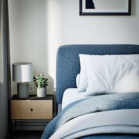 A bed with a blue headboard and white pillows is in the foreground with a nightstand and lamp beside it.