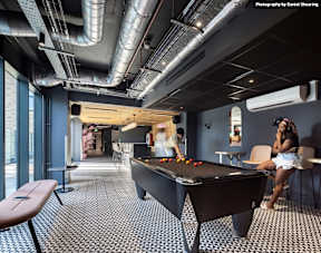 A pool table in a room with a person sitting on a chair.