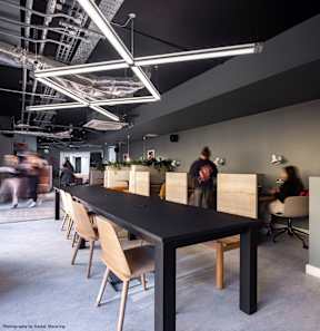 A long black table with tan chairs in a room with people walking in the background.