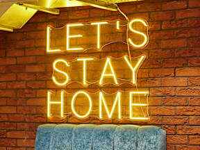 A neon sign that says "Let's Stay Home" is hanging on a brick wall.