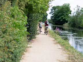 Two people riding bikes on a path next to a river at The Woodlands Apartments, Milton Keynes, MK5 6GS