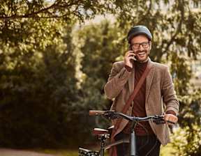 A man in a brown coat and hat is talking on a phone while holding a bicycle at Origins Apartments, Cambridge, CB3 0UX