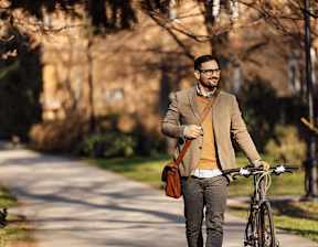 A man in a brown jacket and glasses is walking with a bicycle and holding a brown bag at Origins Apartments, Cambridge, CB3 0UX