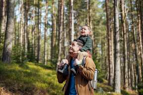 A man is carrying a child on his back while holding a camera in a forest at The Woodlands Apartments, Milton Keynes