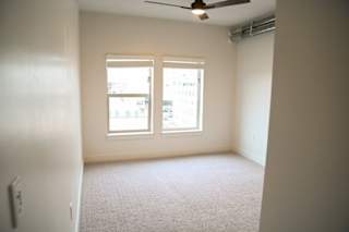A room with a carpeted floor, a ceiling fan, and a window with a view of a building outside.