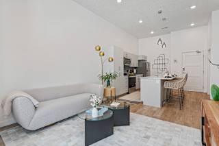 A modern living room with a grey sofa, a glass coffee table, and a kitchen area in the background.