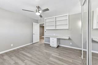 A bedroom with a ceiling fan and a built-in desk with drawers in the middle and shelving above. Vinyl plank flooring and mirrored closet doors.