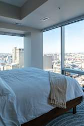 A bed with a white blanket is in front of a window with a view of a cityscape.