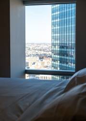 A bed with white sheets sits in front of a window with a view of a cityscape.