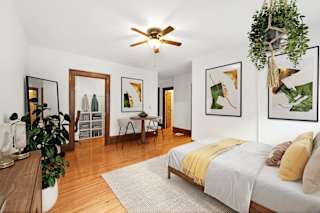 A bedroom with a bed, a ceiling fan, and a dining area in the background.