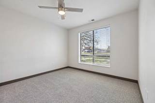 Empty apartment bedroom with neutral carpeting, a ceiling fan, and a large window with blinds overlooking the street.