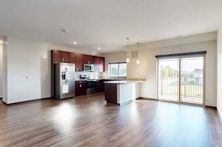 A living room with hardwood flooring, next to an open modern kitchen with dark wooden cabinets, and a sliding glass door to a balcony.
