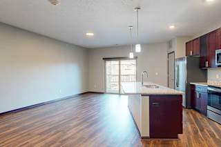 An open-concept living room with hardwood flooring and a sliding glass door to a balcony, that connects to a modern kitchen with a large central island.