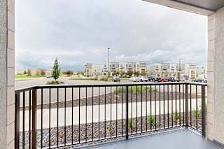 A balcony with a black railing overlooks a parking lot and apartment buildings.