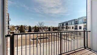 Private apartment balcony with black metal railing overlooking the community courtyard and neighboring residential buildings under a clear blue sky.