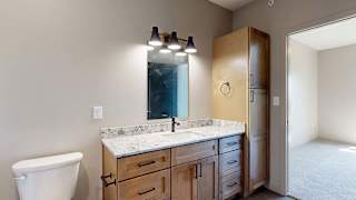 Modern apartment bathroom with a single-sink vanity, wood cabinetry, a stone-look countertop, large mirror, vanity lighting, and an adjoining bedroom visible through the open doorway.