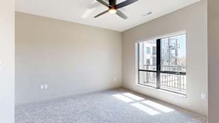 Bright apartment bedroom with soft carpeting, a large window providing natural light, a ceiling fan, neutral finishes, and views of the neighboring community buildings.