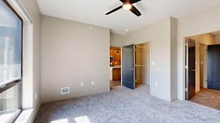 Bright apartment bedroom with soft carpeting, a large window, ceiling fan, neutral walls, and an open doorway leading toward the kitchen and hallway.