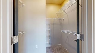 Walk-in apartment closet with carpeted flooring, multiple wire shelving units, and ample storage space for clothing and household items.