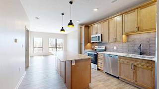 Bright apartment kitchen with wood cabinetry, a large island, tile backsplash, stainless steel appliances, pendant lighting, and an open layout connecting to the living area.