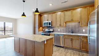 Spacious apartment kitchen with wood cabinetry, a large island, tile backsplash, stainless steel appliances, pendant lighting, and an open layout with adjacent living space and large windows.