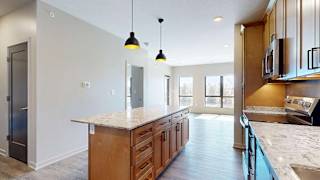 Spacious apartment kitchen with a large island, wood cabinetry, stainless steel appliances, pendant lighting, stone-look countertops, and an open layout flowing into the bright living area.