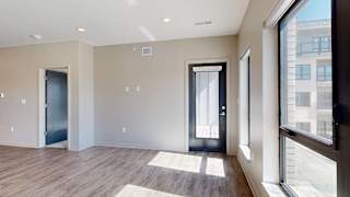 Bright open-concept apartment living area with wood-style flooring, large windows, abundant natural light, and a glass door leading to a private balcony.