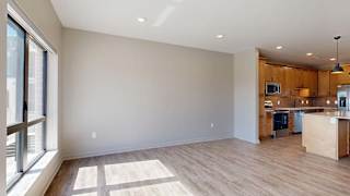 Bright open-concept apartment living room and kitchen with wood-style flooring, large windows, natural light, wood cabinetry, a kitchen island, and stainless steel appliances.