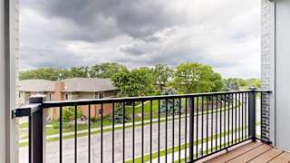 Private apartment balcony with black metal railing and views of trees and neighboring homes under a cloudy sky.