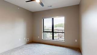 Bright apartment bedroom with carpeted flooring, a large window, and a ceiling fan.