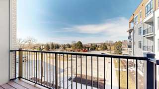 Private apartment balcony with black metal railing overlooking a courtyard, neighboring buildings, trees, and open blue sky.