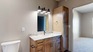 Modern apartment bathroom with a quartz-style vanity, wood cabinetry, contemporary lighting, and a built-in linen cabinet, with the adjacent bedroom visible through the doorway.